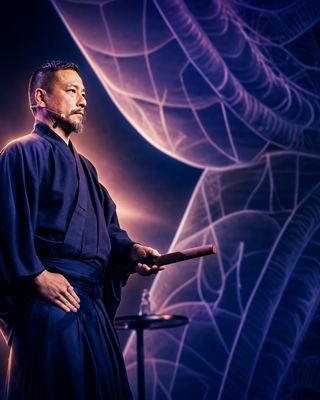A speaker in traditional attire presents on stage at the HOLOLIFE Summit, set against a vivid backdrop of biohacking visuals.